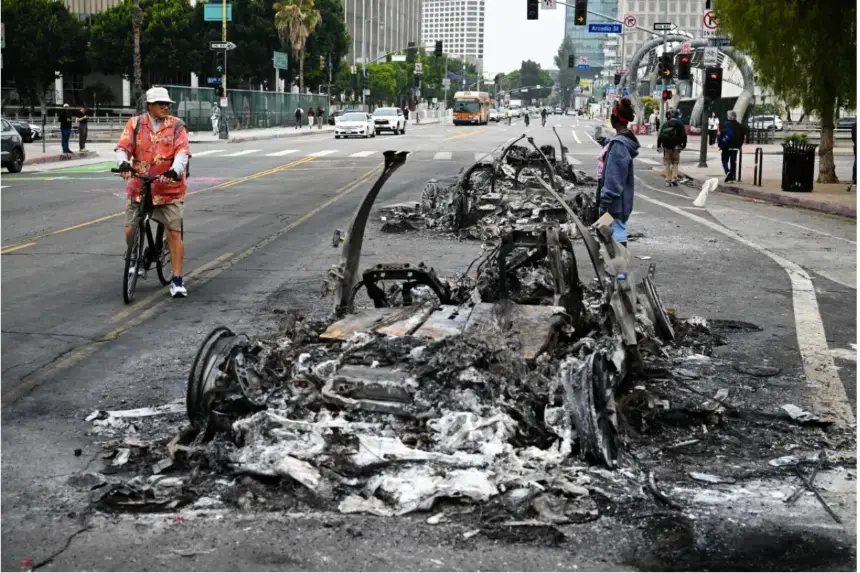 La gente observa los restos carbonizados de un vehículo Waymo incendiado en la calle tras una noche de protestas en respuesta a las operaciones federales de inmigración en Los Ángeles, el 9 de junio de 2025. (Ronaldo Schemidt/AFP vía Getty Images)