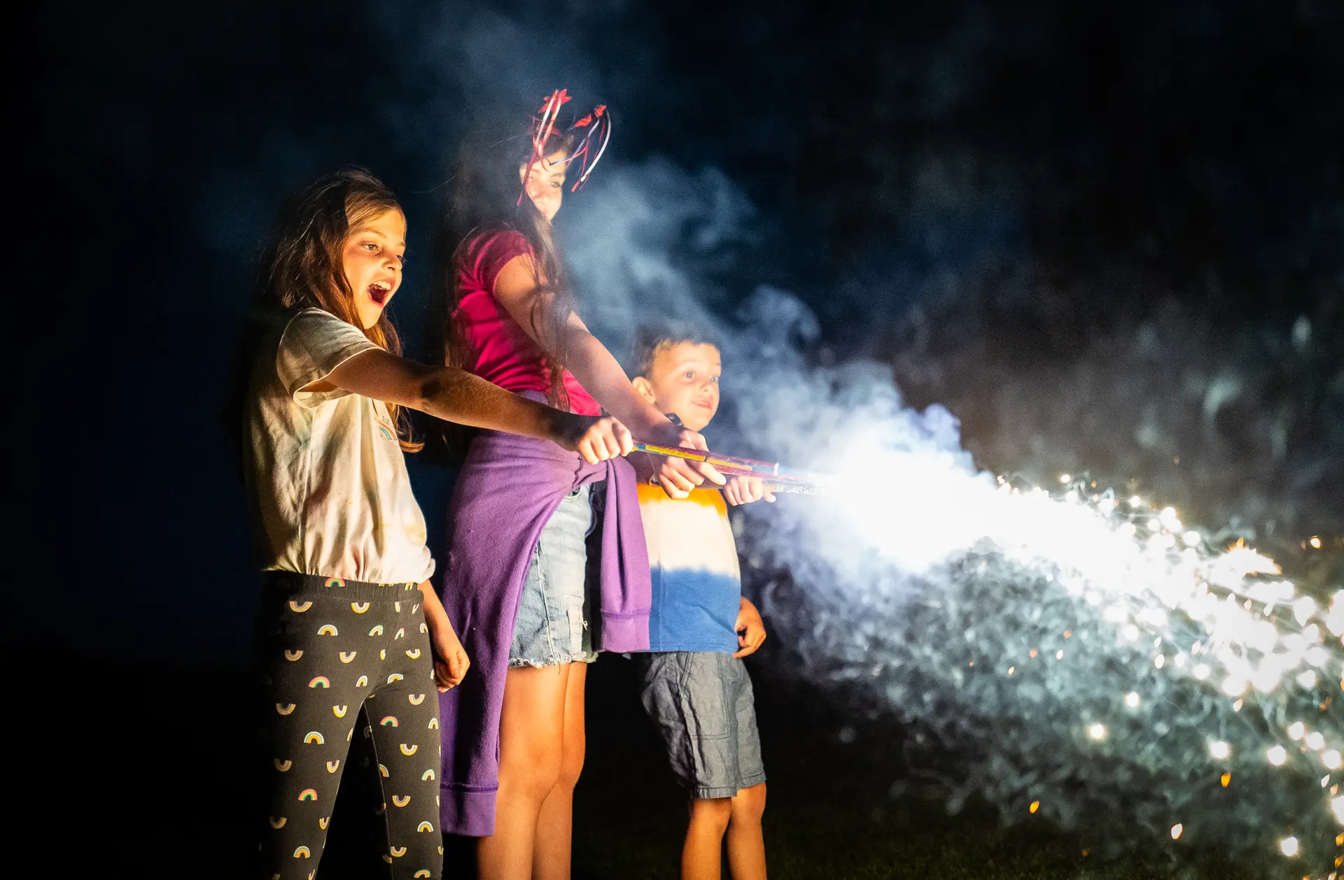 Los hermanos Aria Equils de 6 años (izq.), Evie Equils de 11 años (centro) y Brody Equils de 6 años (der.), juegan con bengalas en Temecula, California, el 4 de julio de 2025. (John Fredricks/The Epoch Times)