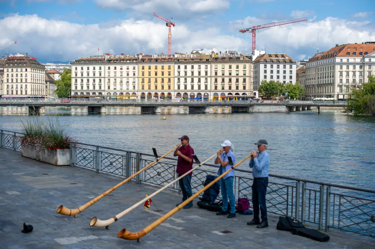 Los edificios coronados con los logotipos de Vacheron Constantin, Zenith, Baume & Mercier y Breitling son visibles desde el lago Lemán, con trompetistas tocando en primer plano en la orilla opuesta, en Ginebra, Suiza, el 2 de agosto de 2025. (Robert Hradil/Getty Images).