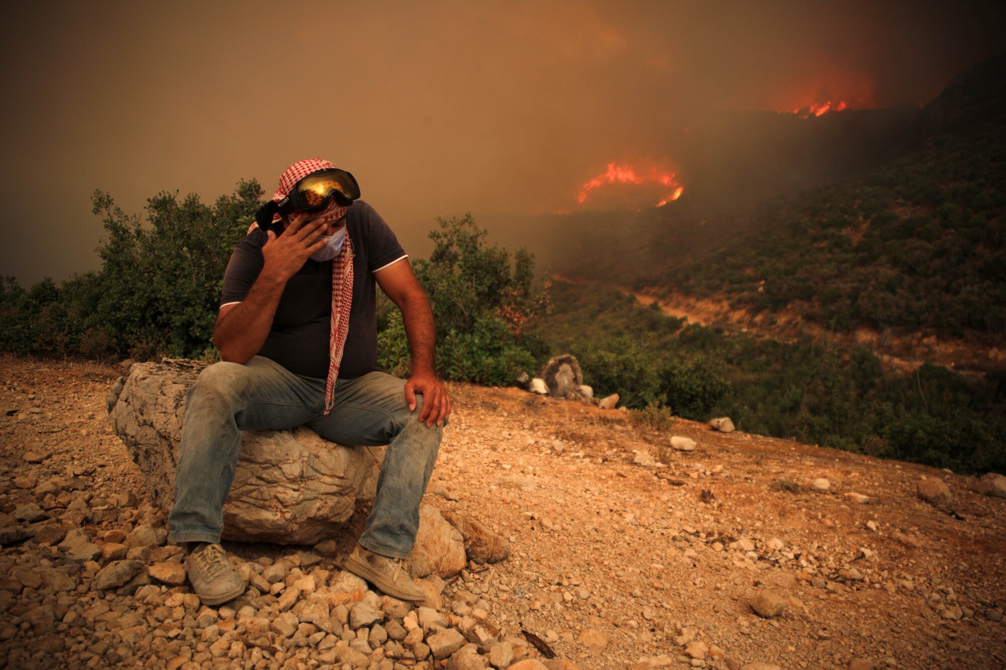 Un residente con una mascarilla sentado en una roca mientras un incendio forestal arrasa una colina detrás de él, en la zona rural occidental de Hama, Siria, el 15 de agosto de 2025. (Abdulaziz Ketaz/AFP a través de Getty Images).