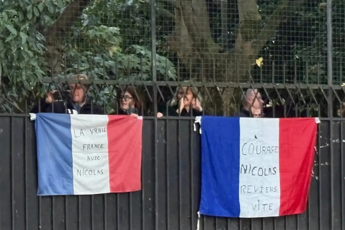 La gente se reúne detrás de banderas francesas con una inscripción que dice “Ánimo, Nicolas, vuelve pronto” y otra que dice “La verdadera Francia con Nicolas”, frente a la casa del expresidente francés Nicolas Sarkozy en París, el 21 de octubre de 2025. (Foto AP/Masha Macpherson)