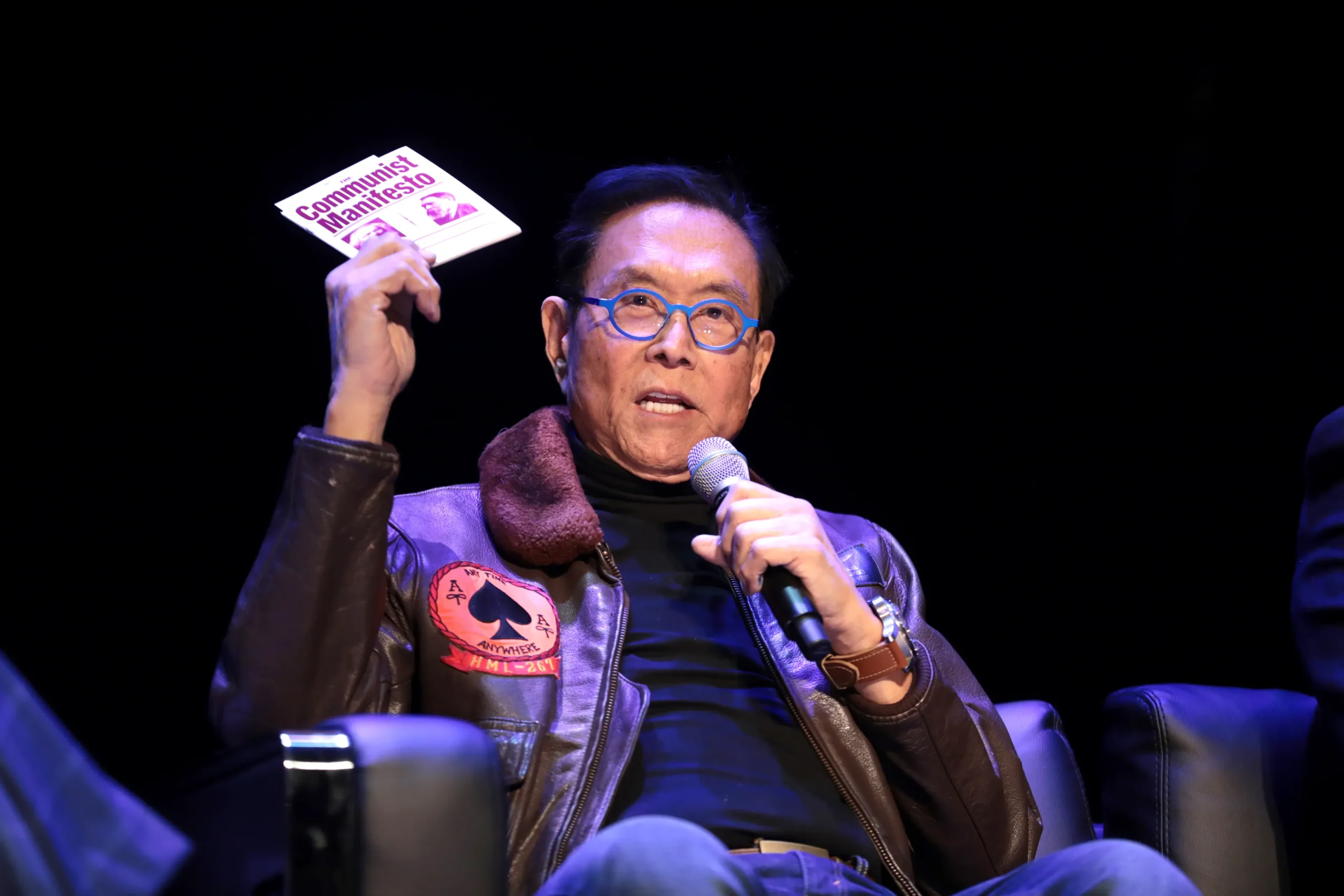 <em>Robert Kiyosaki sostiene un ejemplar de "El Manifiesto Comunista" mientras habla con los asistentes a un evento de la Universidad Estatal de Arizona en Tempe, Arizona, el 8 de febrero de 2023. Kiyosaki dijo que su profesor de economía de la universidad lo instó a leer el libro de Karl Marx para "conocer a su enemigo". (Gage Skidmore/Flickr)</em>